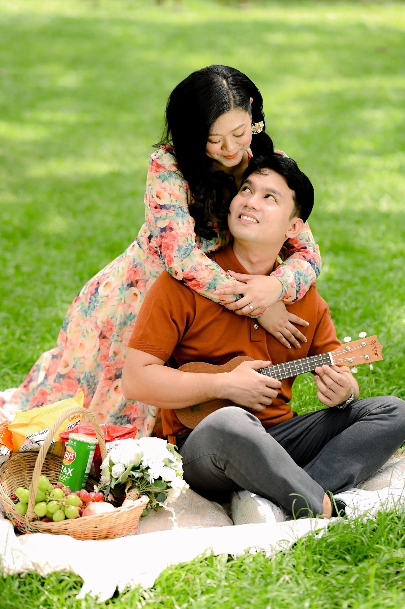 Chie and Cath at a picnic on the grass, wrapped in an embrace, with a ukulele and a basket of fruit and flowers.